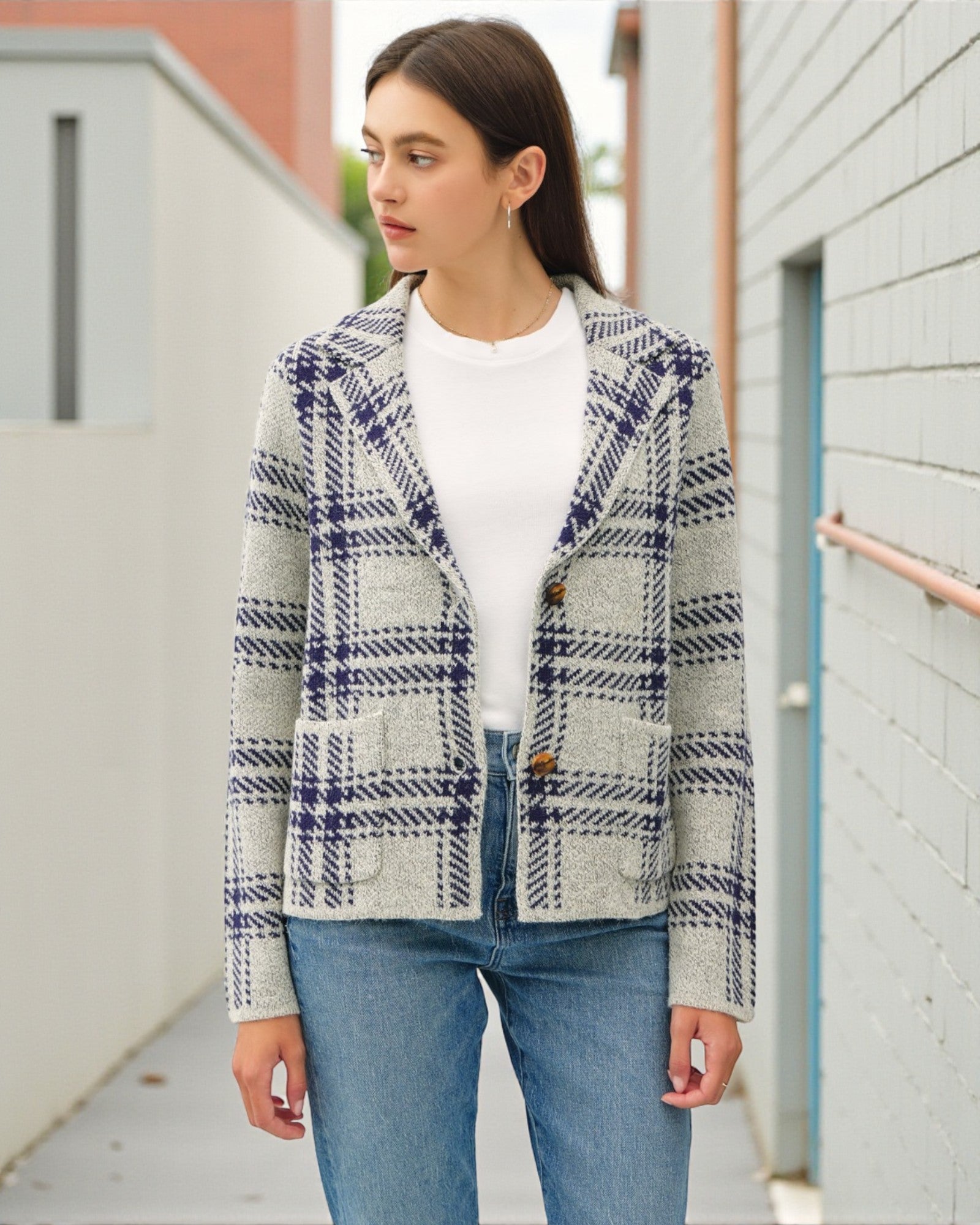 Woman wearing a plaid jacket over a white shirt and blue jeans, standing against a light-colored wall.