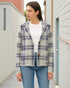 Woman wearing a plaid jacket over a white shirt and blue jeans, standing against a light-colored wall.