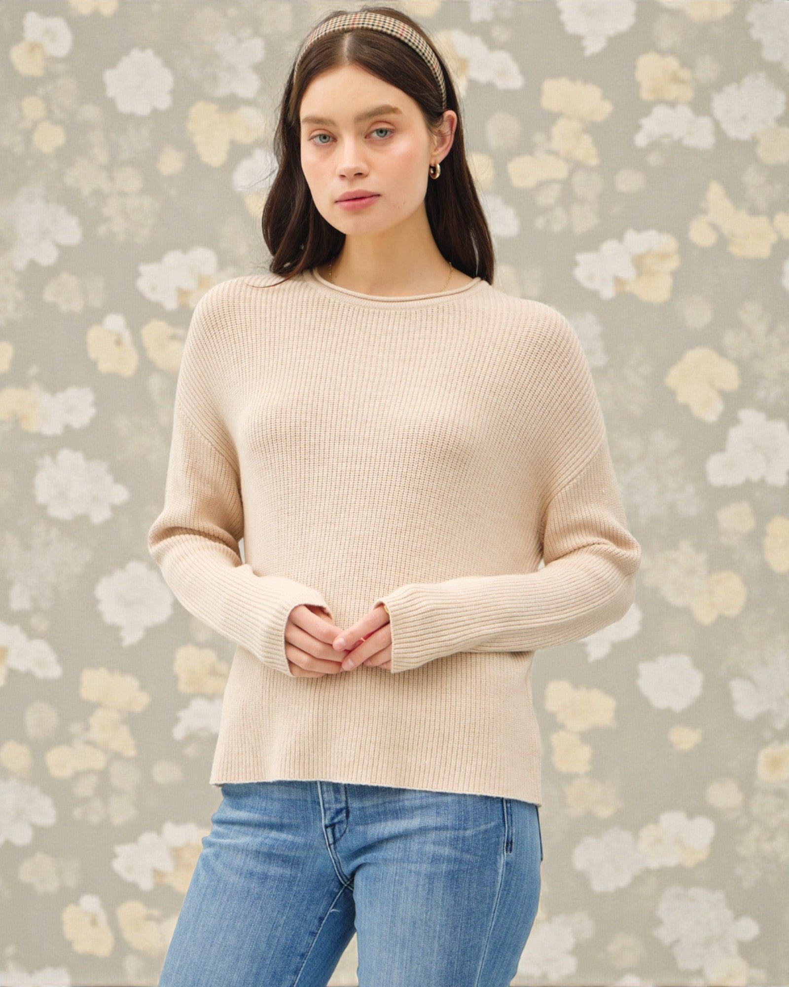 Woman wearing a beige sweater and blue jeans against a floral-patterned background