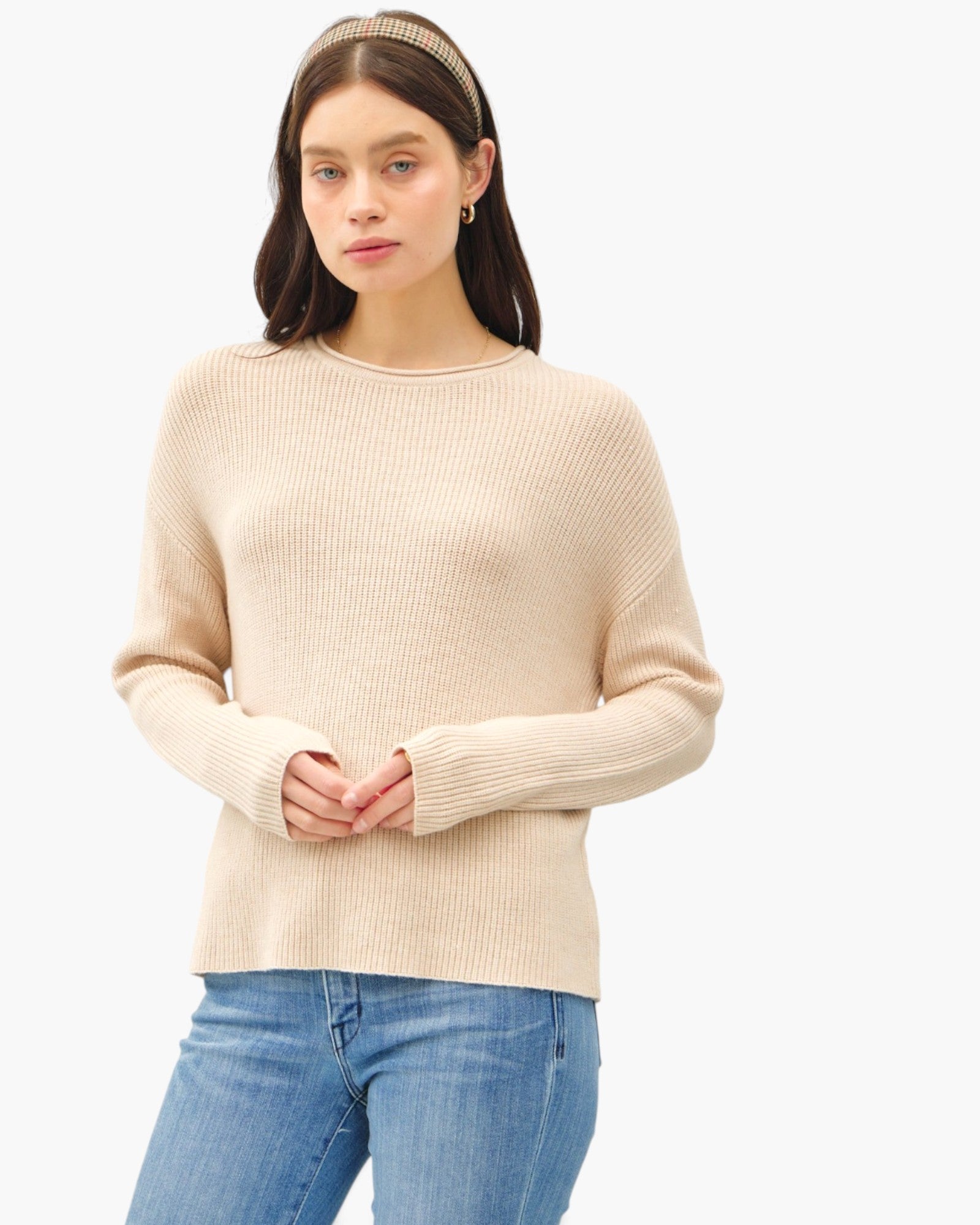 A woman wearing a beige ribbed knit sweater and blue jeans, standing against a plain white studio background.