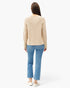 A woman viewed from behind wearing a light beige knit sweater, blue cropped jeans, and white slip-on shoes against a plain white studio background.