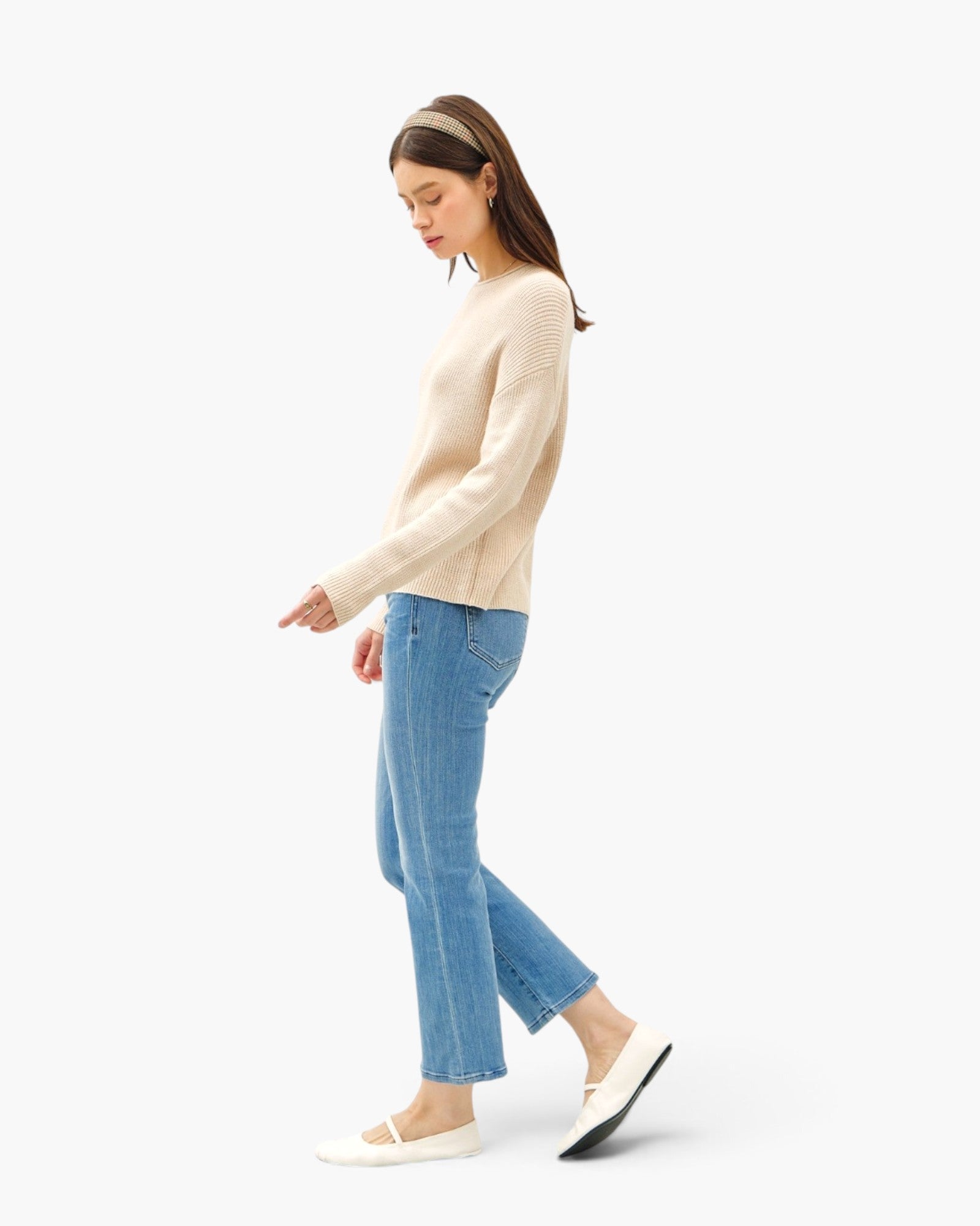 A woman in a side profile wearing a beige knit sweater, light blue cropped jeans, and white Mary Jane flats, set against a plain white studio background.