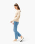 A woman in a side profile wearing a beige knit sweater, light blue cropped jeans, and white Mary Jane flats, set against a plain white studio background.