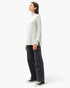 A woman stands in profile wearing a light gray mock-neck knit sweater, dark wide-leg jeans, and black low-top sneakers against a plain light gray studio background.