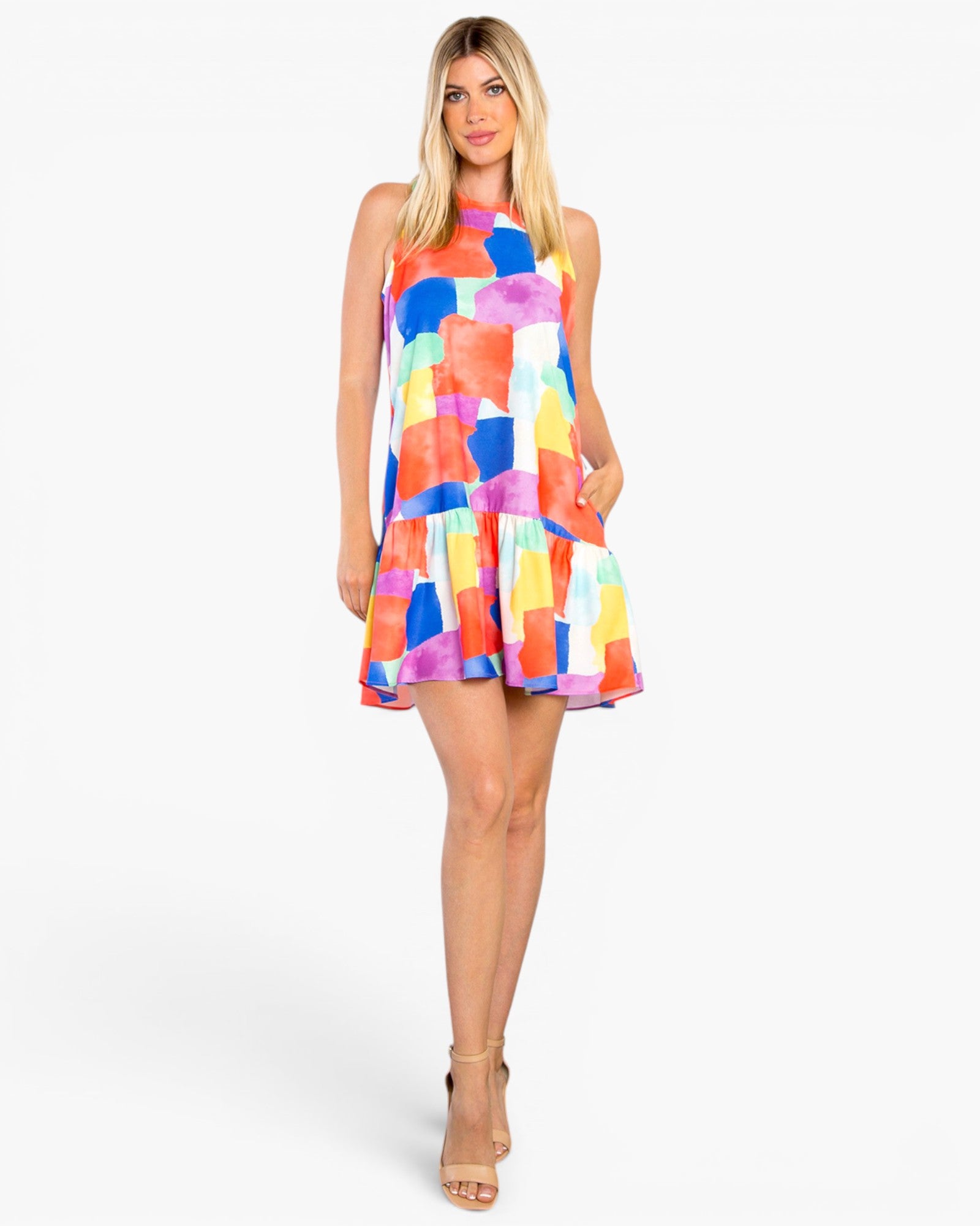A woman modeling a sleeveless tiered mini dress with a vibrant multicolor abstract watercolor pattern in shades of blue, pink, orange, yellow, and purple against a plain white studio background.