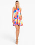 A woman modeling a sleeveless tiered mini dress with a vibrant multicolor abstract watercolor pattern in shades of blue, pink, orange, yellow, and purple against a plain white studio background.