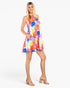 A woman modeling a sleeveless tiered mini dress with a vibrant multicolor abstract geometric print, paired with beige block-heel sandals, against a plain white studio background.
