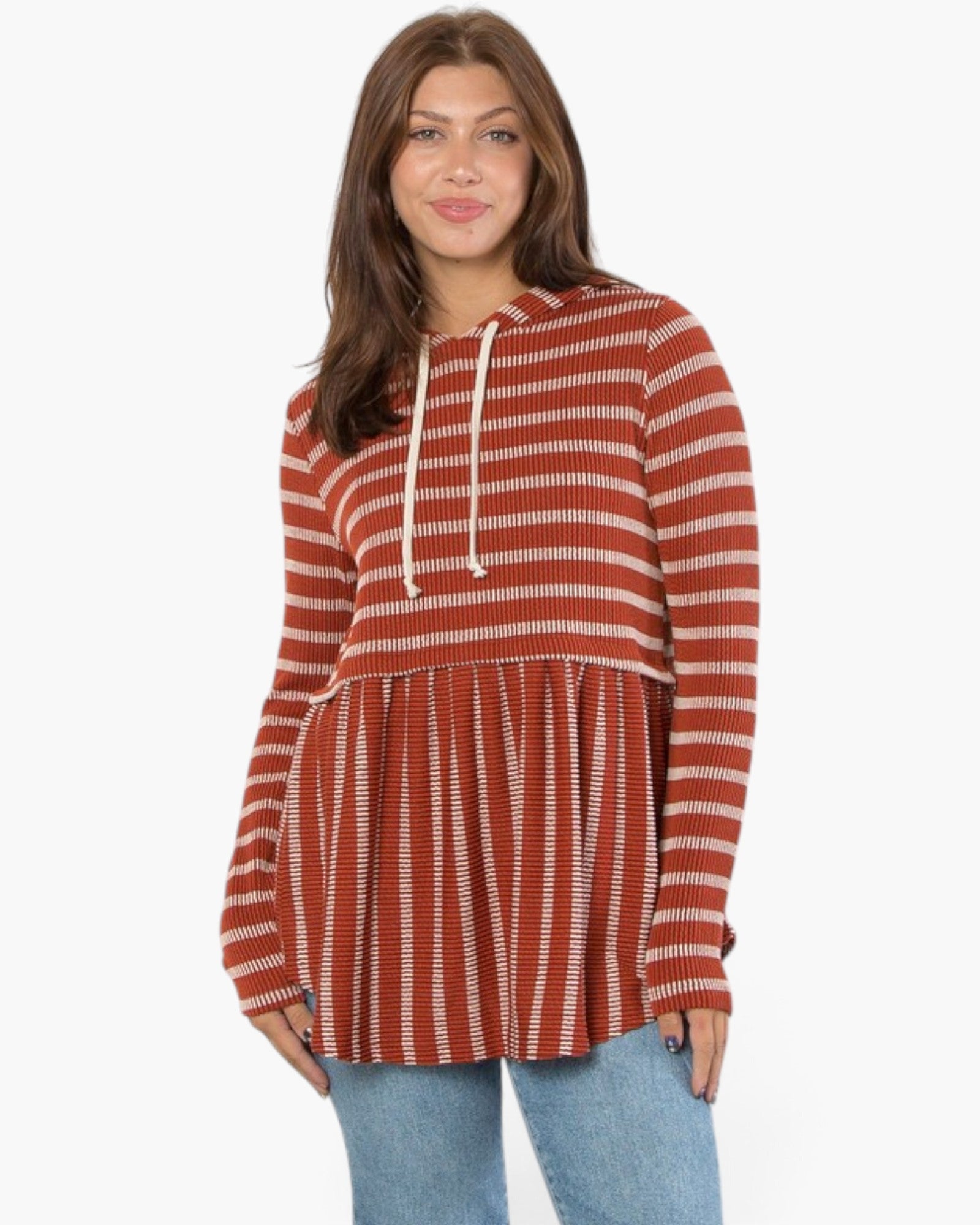 A woman wearing a rust-orange and white horizontally striped hooded long-sleeve tunic top with a flared peplum hem, paired with light blue jeans, against a plain white background.