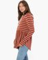 A woman wearing a rust-orange and white striped long-sleeve knit top with a drawstring neckline and a flared peplum hem, paired with light blue jeans, against a plain white background.