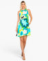 A woman modeling a sleeveless halter-neck mini dress with a bold retro floral pattern in turquoise, lime green, yellow, white, and black, set against a plain light gray studio background.