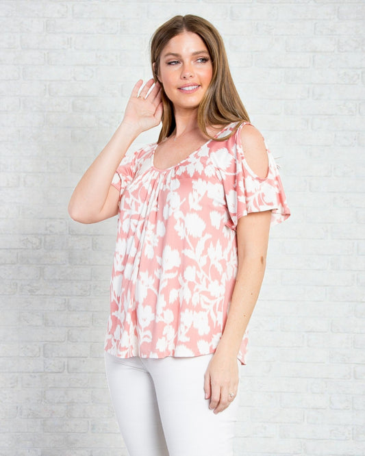 Woman wearing a pink floral blouse with cold shoulder design against a light gray brick wall.