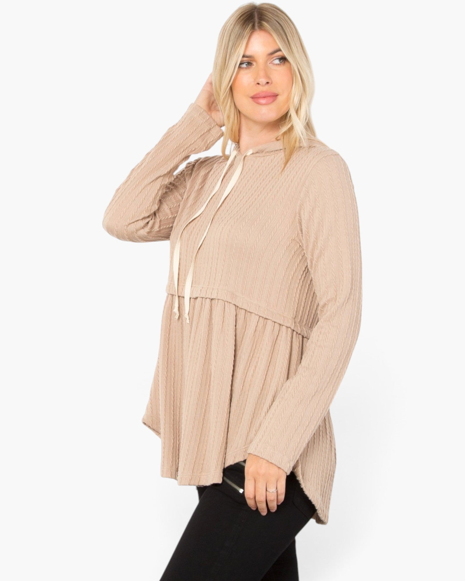 A woman modeling a beige ribbed knit long-sleeve hooded tunic top with cream drawstrings, paired with black pants, against a plain white studio background.