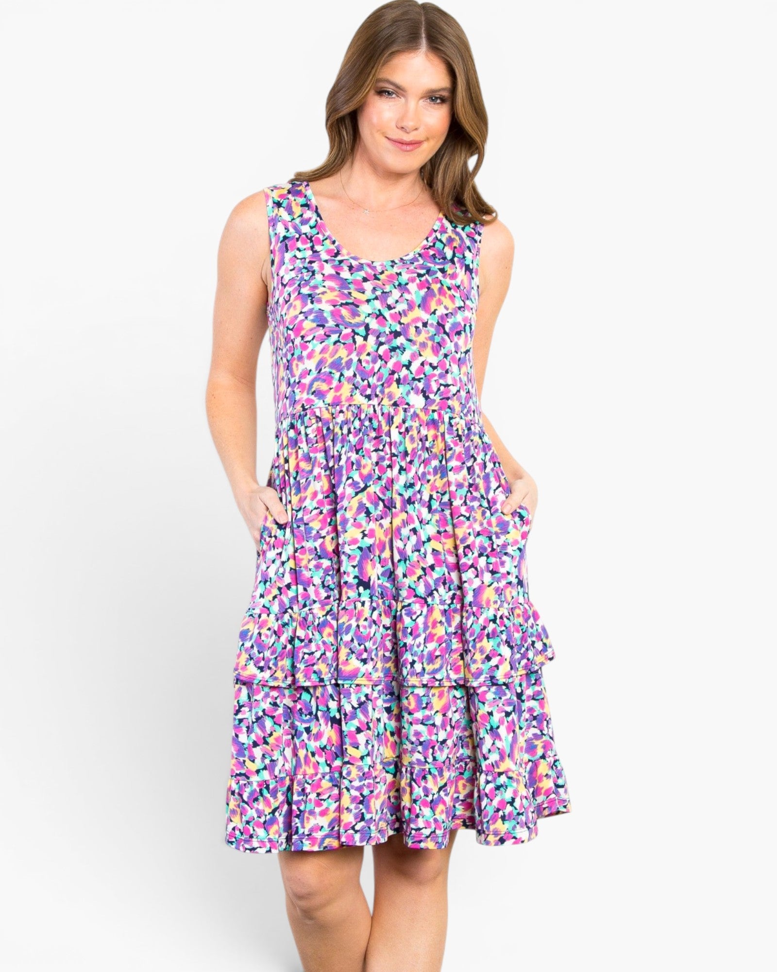 A woman modeling a sleeveless, tiered A-line dress with a vibrant multicolor abstract print in shades of purple, pink, blue, and yellow against a plain white studio background.