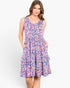 A woman modeling a sleeveless, tiered A-line dress with a vibrant multicolor abstract print in shades of purple, pink, blue, and yellow against a plain white studio background.