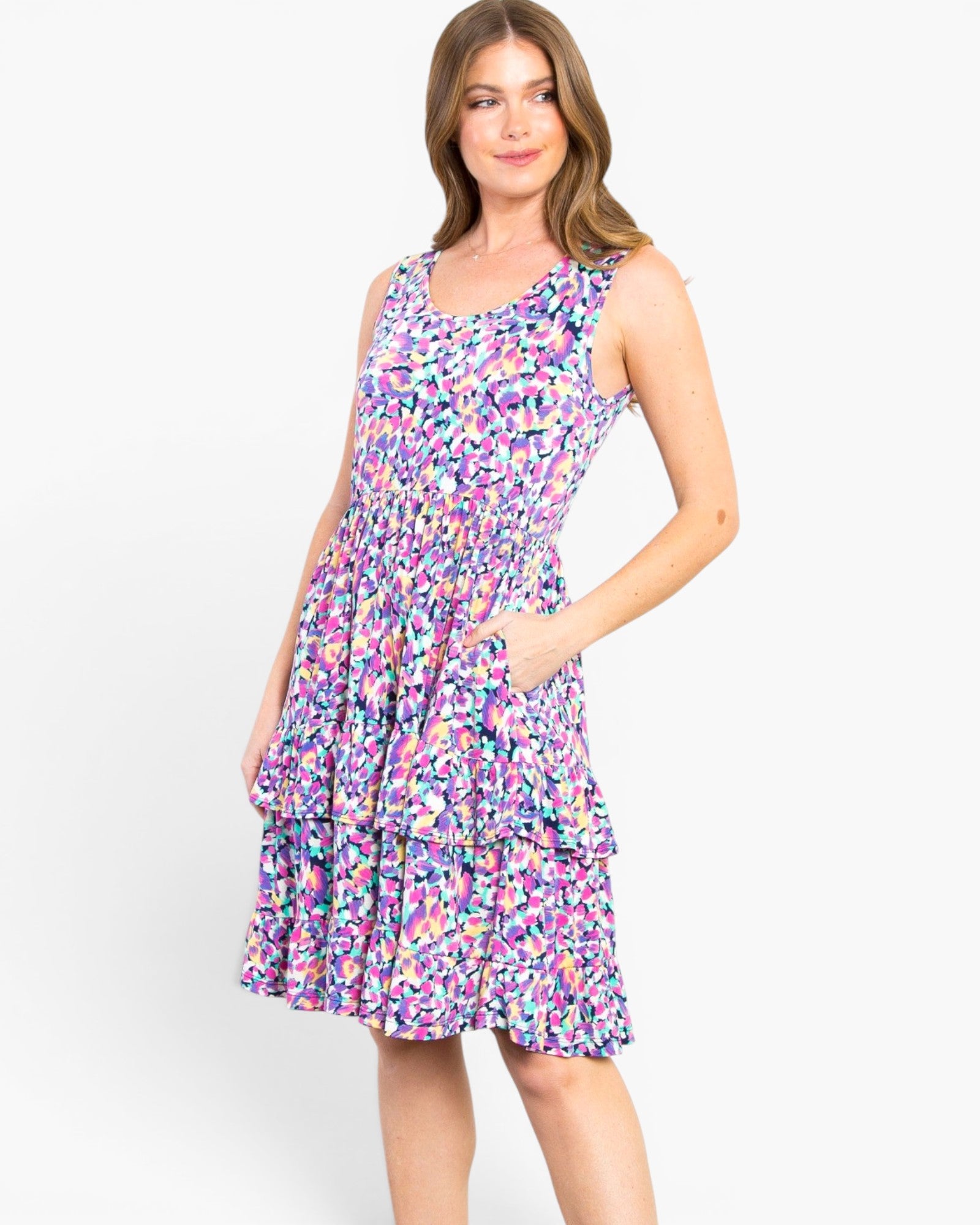 A woman modeling a sleeveless tiered A-line dress with a colorful abstract floral print in purple, pink, blue, and white, set against a plain white studio background.