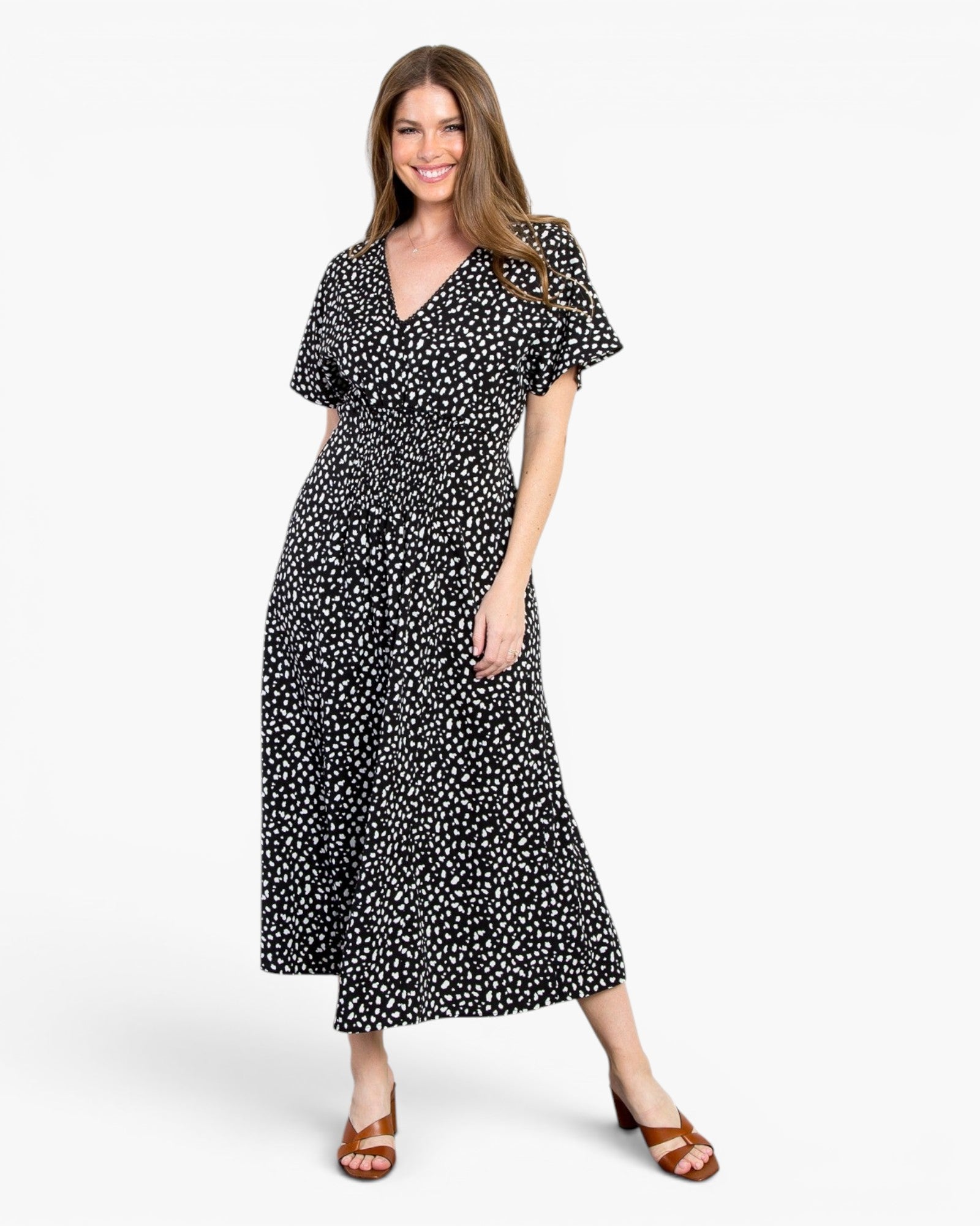 A woman modeling a black midi dress with a white ditsy floral print, featuring short flutter sleeves and a V-neckline, paired with brown heeled sandals against a plain white studio background.