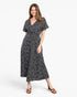 A woman modeling a black midi dress with a white ditsy floral print, featuring short flutter sleeves and a V-neckline, paired with brown heeled sandals against a plain white studio background.