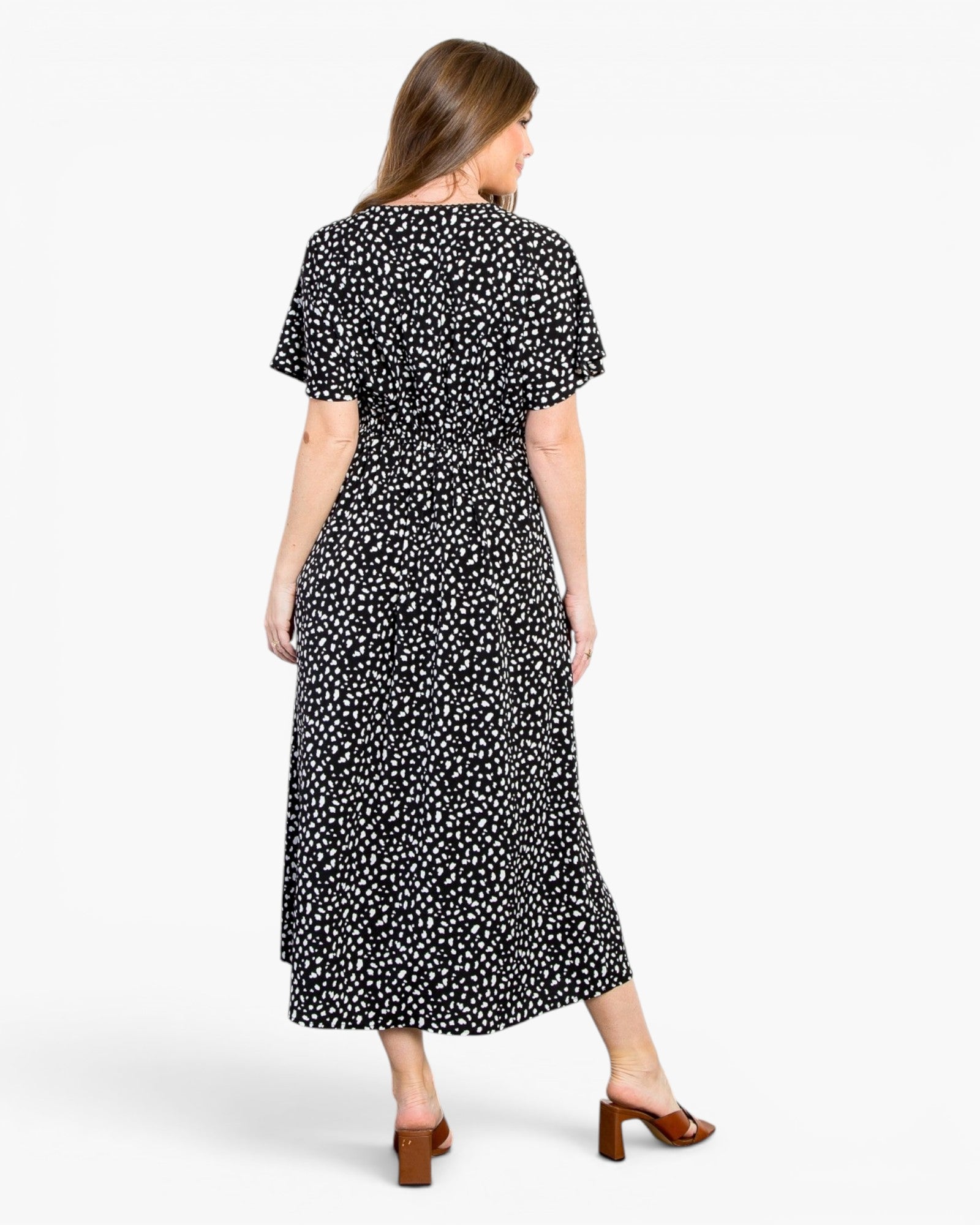 A woman viewed from the back wearing a short-sleeve midi dress with a black base and small white floral print, paired with brown heeled sandals against a light gray studio background.