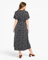 A woman viewed from the back wearing a short-sleeve midi dress with a black base and small white floral print, paired with brown heeled sandals against a light gray studio background.