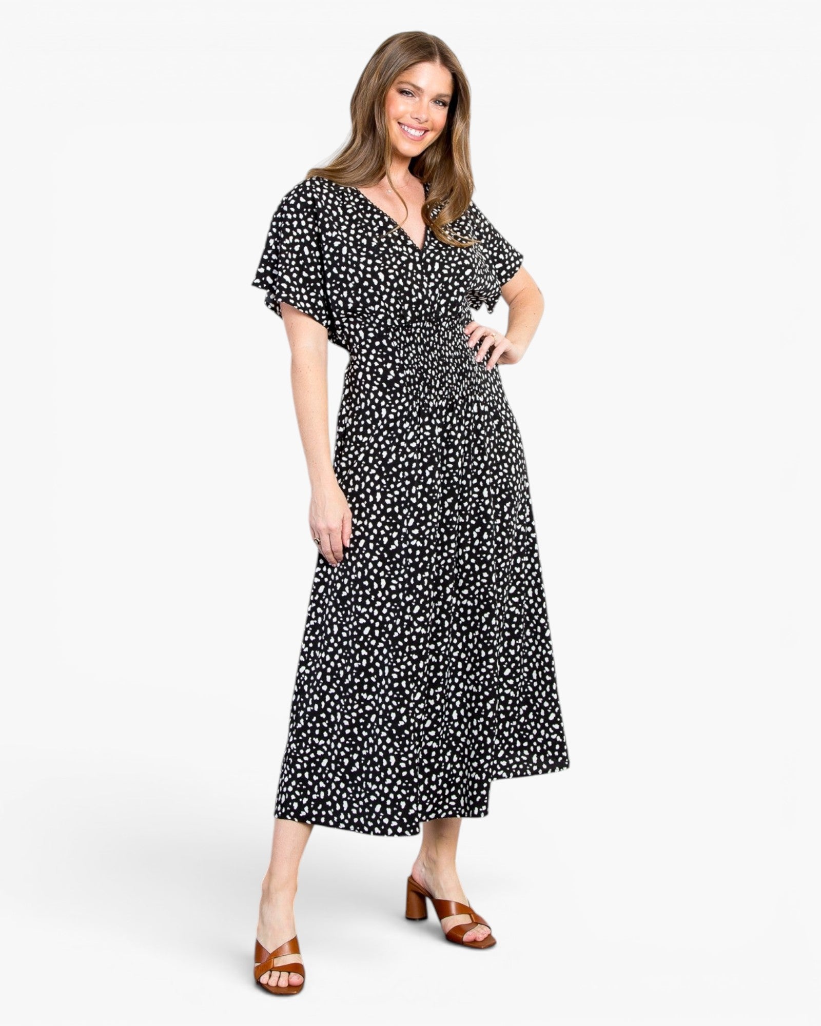 A woman modeling a black midi dress with a white ditsy floral print, featuring flutter sleeves and a V-neckline, paired with brown strappy heeled sandals against a plain white studio background.