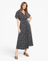 A woman modeling a black midi dress with a white ditsy floral print, featuring flutter sleeves and a V-neckline, paired with brown strappy heeled sandals against a plain white studio background.