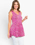 A woman modeling a sleeveless V-neck peplum top with a vibrant pink, black, and white abstract pattern, paired with white pants, against a plain white studio background.