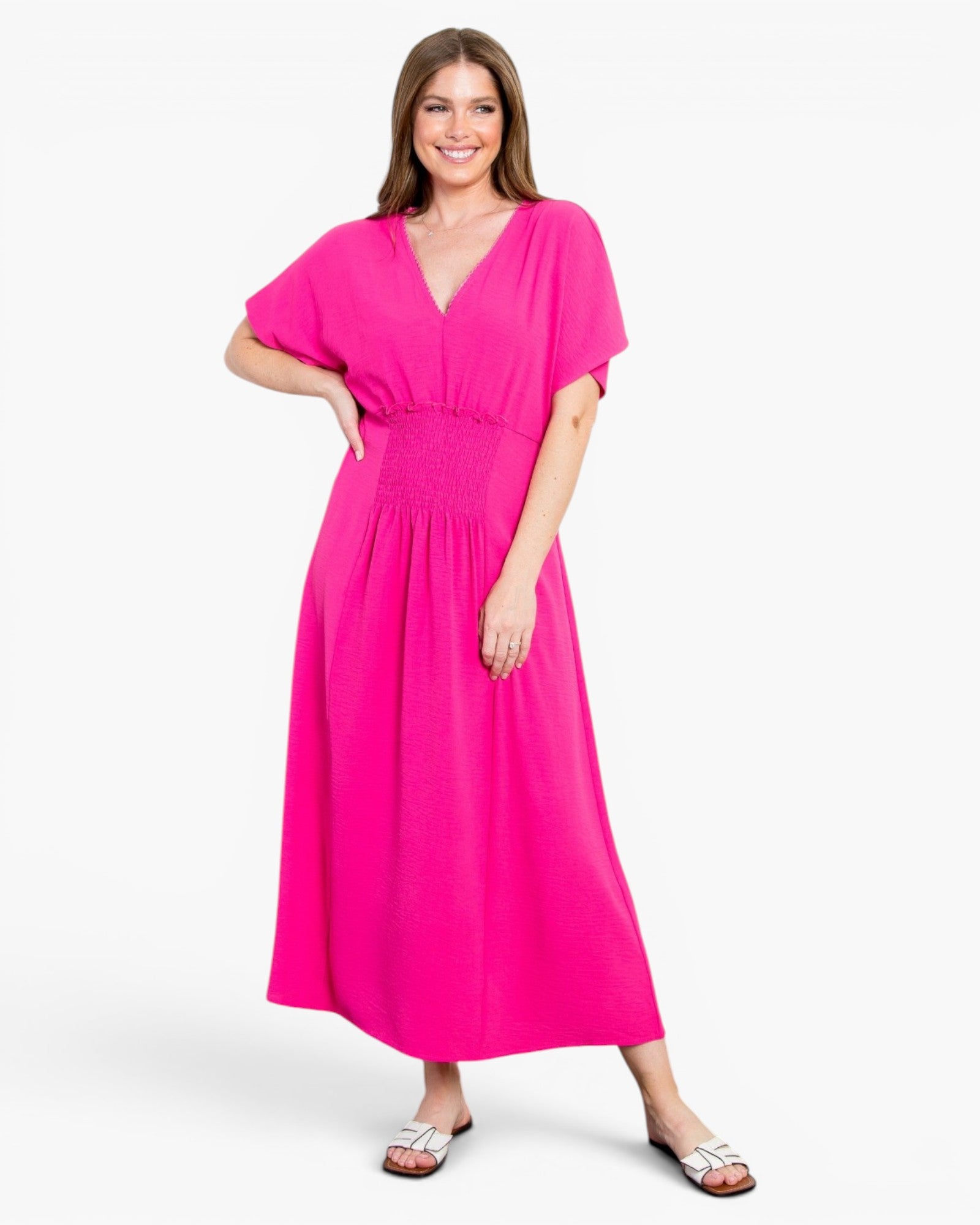 A woman modeling a bright pink midi dress with a V-neckline, short flutter sleeves, and a smocked waistband, paired with white slide sandals, against a light gray studio background.
