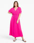 A woman modeling a bright pink midi dress with a V-neckline, short flutter sleeves, and a smocked waistband, paired with white slide sandals, against a light gray studio background.