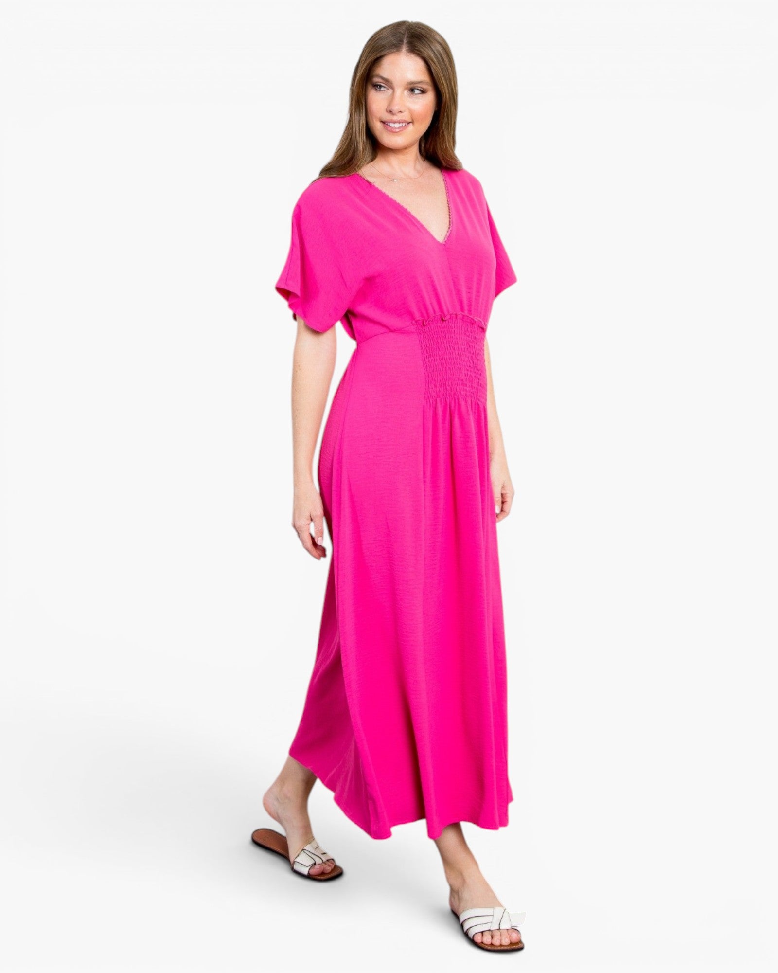 A woman modeling a bright pink maxi dress with short flutter sleeves, a V-neckline, and a smocked waistband, paired with brown slide sandals against a light gray studio background.