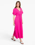 A woman modeling a bright pink maxi dress with short flutter sleeves, a V-neckline, and a smocked waistband, paired with brown slide sandals against a light gray studio background.