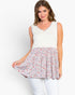 A woman modeling a sleeveless V-neck top with a solid cream upper section and a flared floral skirt in white, pink, and blue, paired with white pants against a light gray studio background.