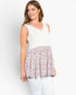 A woman modeling a sleeveless top with a solid cream upper bodice and a flared floral skirt in pink, red, and blue tones, paired with white pants against a plain white background.