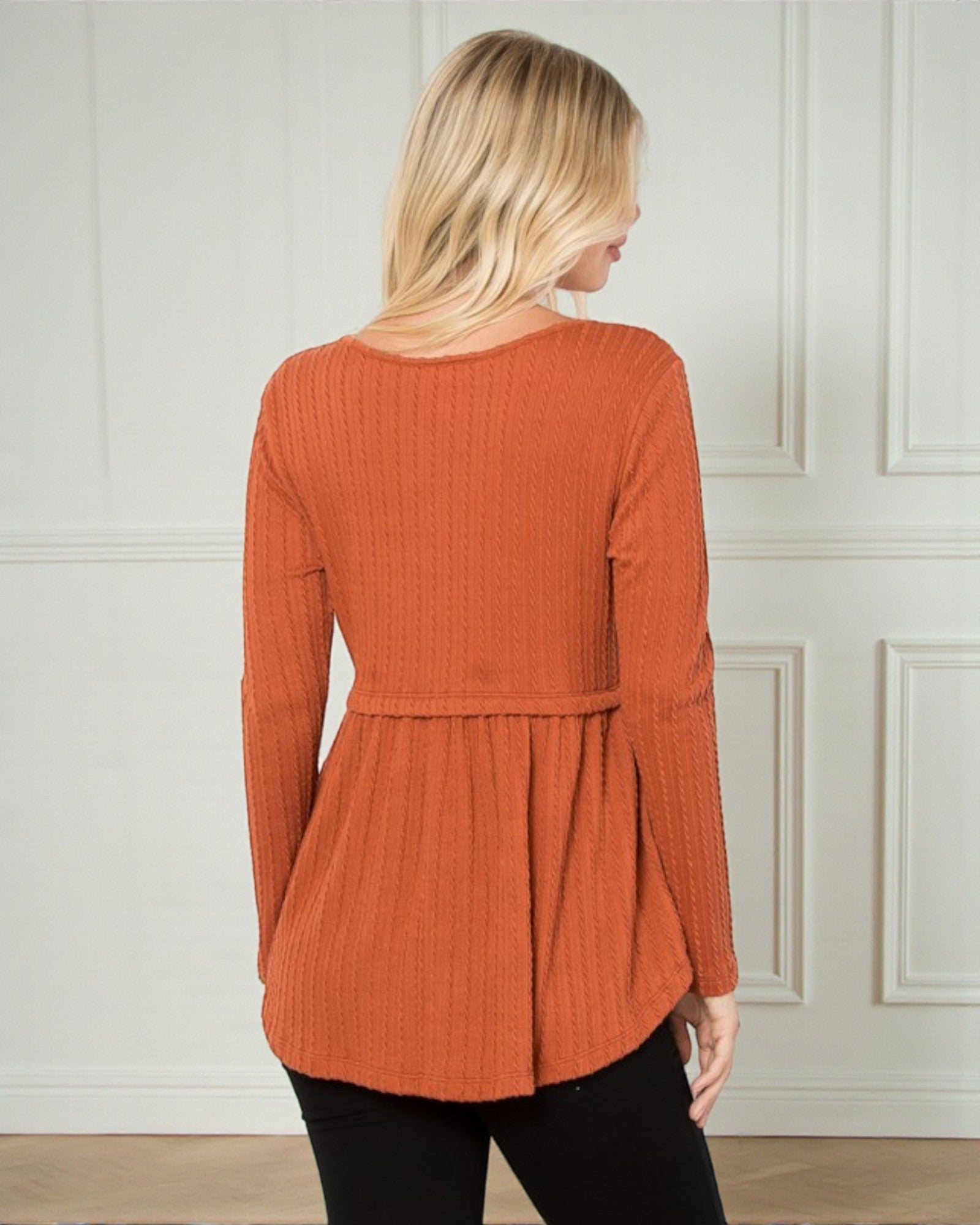 Woman wearing an orange ribbed long-sleeve top against a white paneled wall.