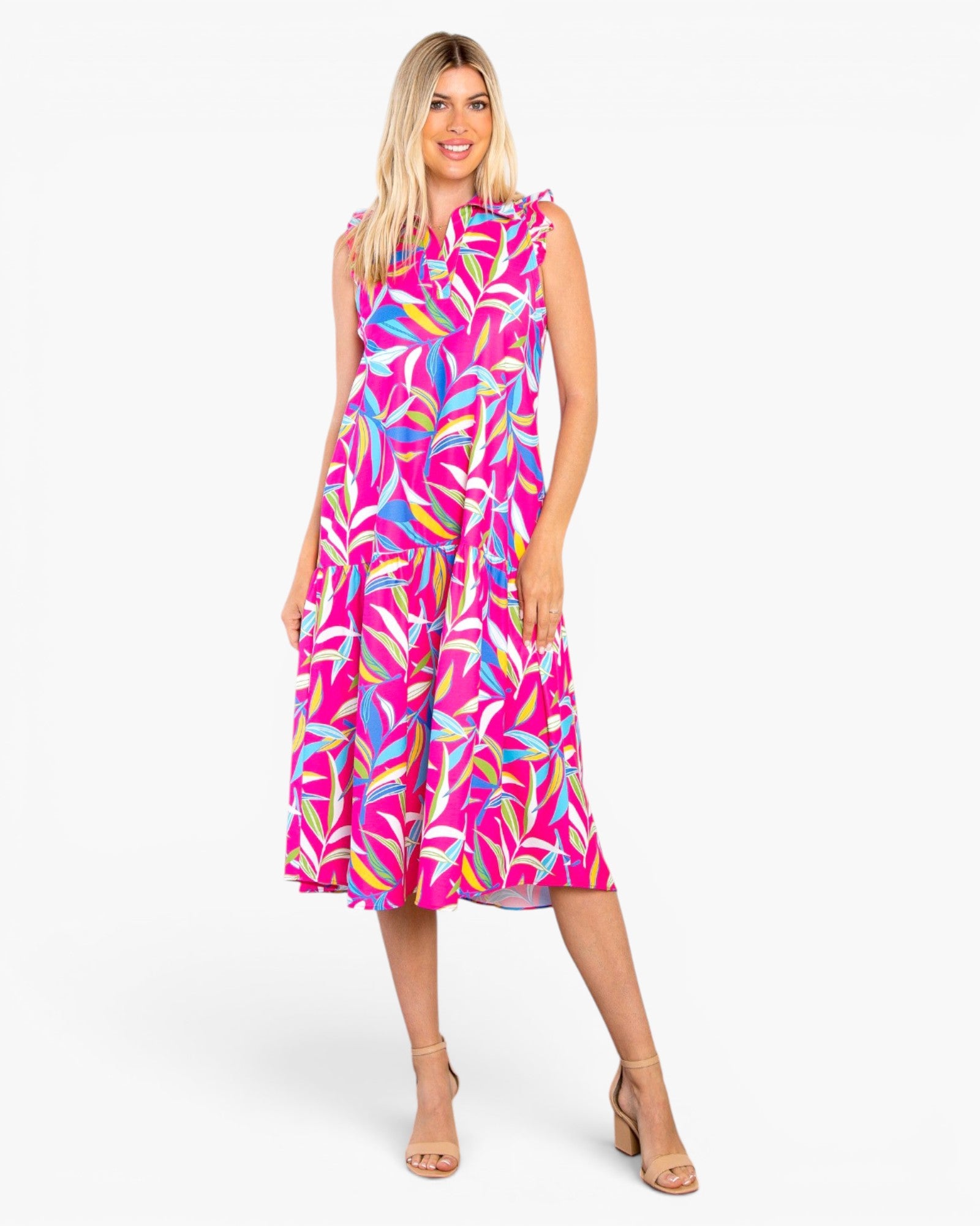 A woman modeling a sleeveless midi dress with a bright pink and multicolor tropical leaf print, paired with beige high-heeled sandals, against a plain white studio background.