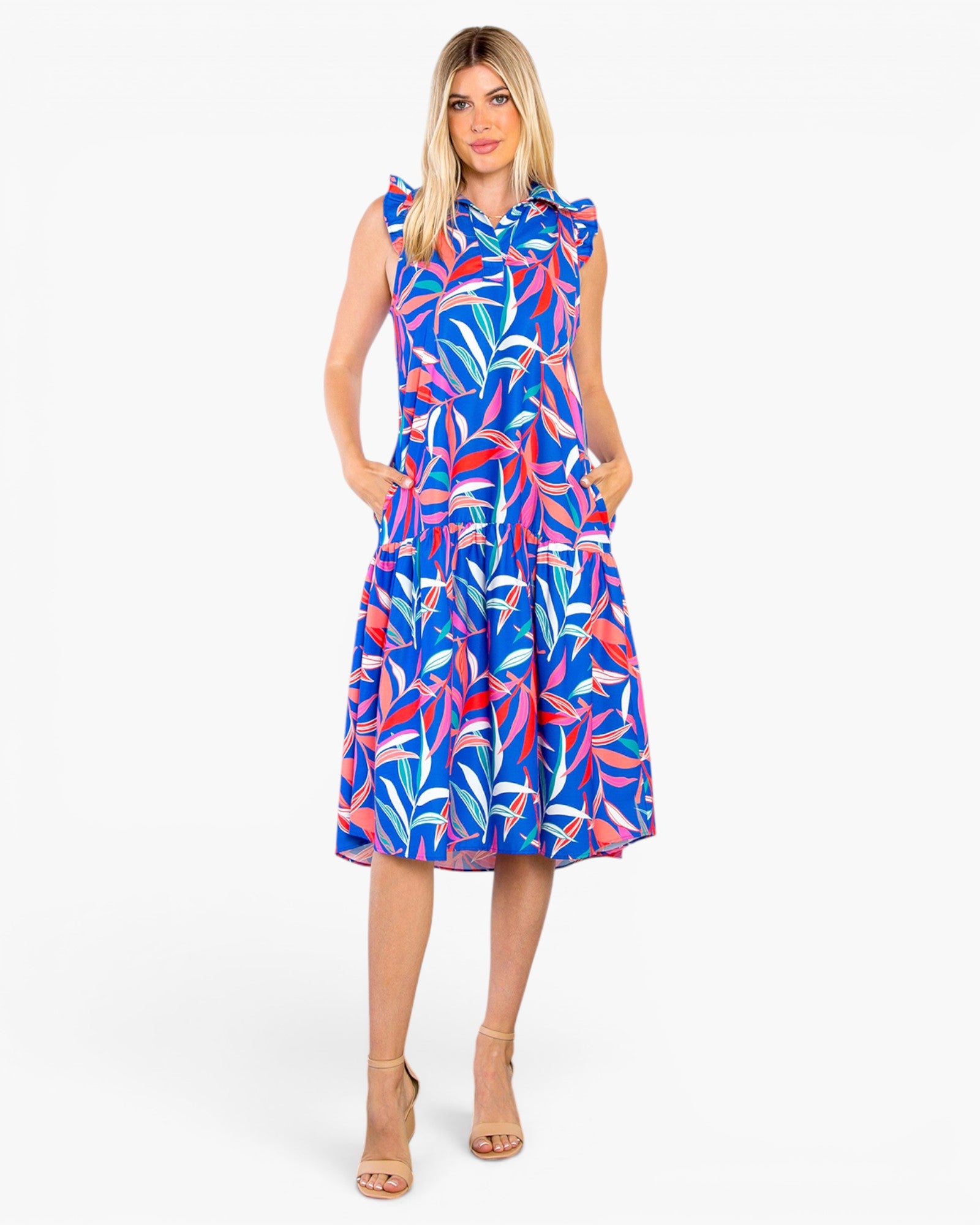 A woman modeling a sleeveless tiered midi dress with a vibrant blue, pink, and white tropical leaf pattern, set against a plain light gray background.
