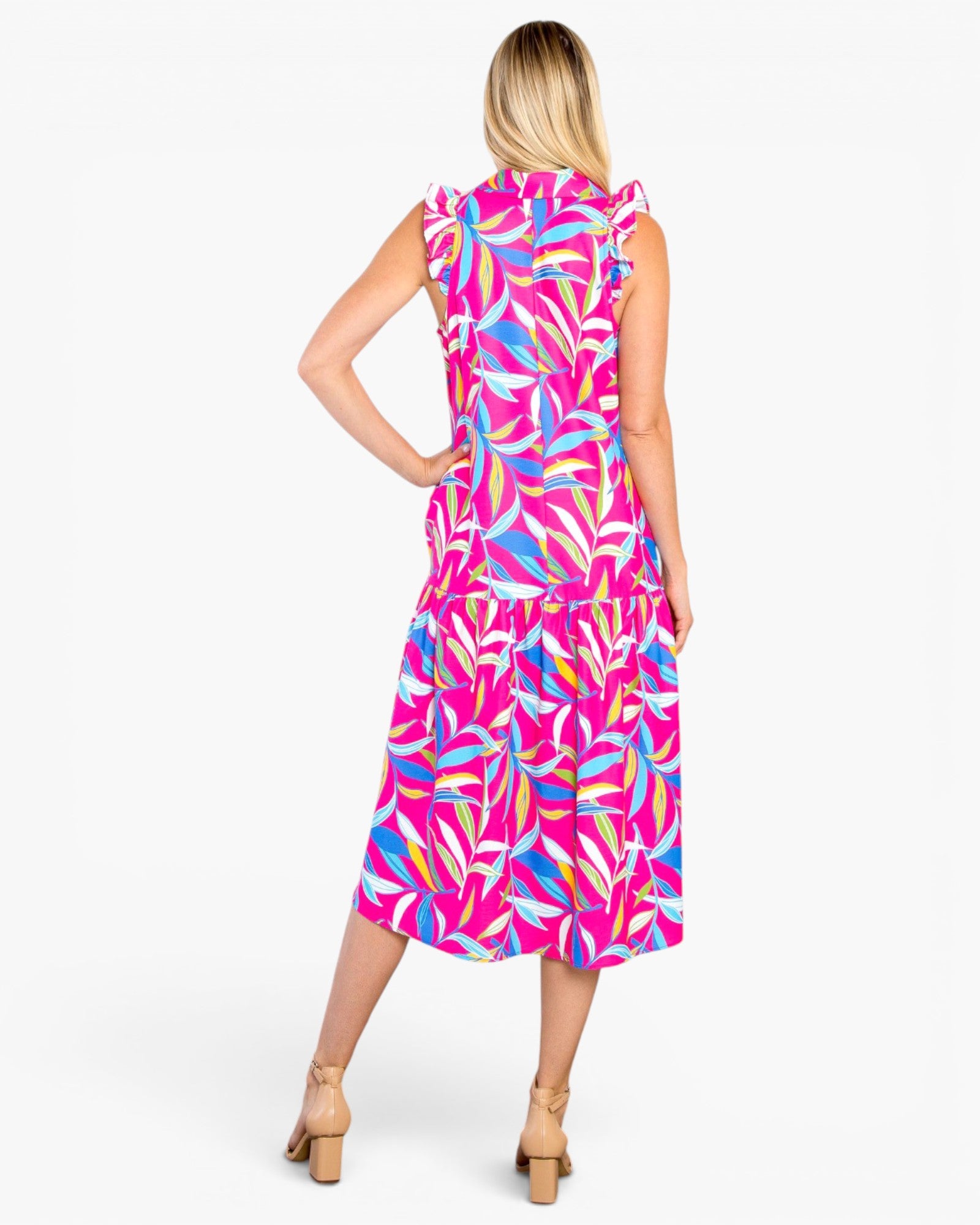 A woman modeling a sleeveless midi dress from the back, featuring a vibrant pink, blue, and yellow tropical leaf print, set against a plain white studio background.