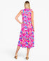 A woman modeling a sleeveless midi dress from the back, featuring a vibrant pink, blue, and yellow tropical leaf print, set against a plain white studio background.