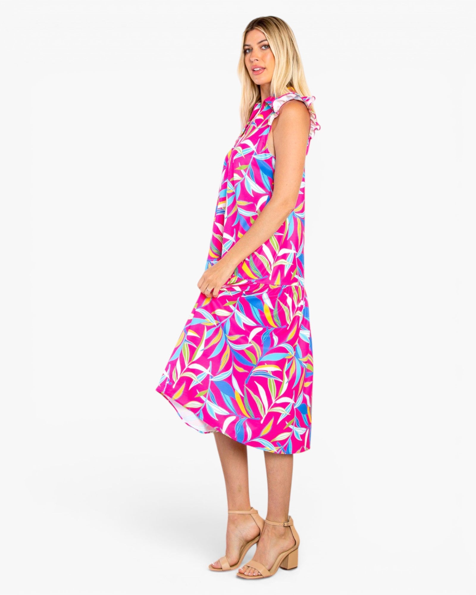 A woman modeling a sleeveless midi dress with a bright pink, blue, and orange tropical leaf print, shown from a side profile against a light gray studio background.