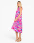 A woman modeling a sleeveless midi dress with a bright pink, blue, and orange tropical leaf print, shown from a side profile against a light gray studio background.