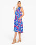 A woman modeling a sleeveless knee-length dress with a vibrant blue, pink, and white tropical leaf pattern, paired with beige block-heel sandals, set against a plain white studio background.