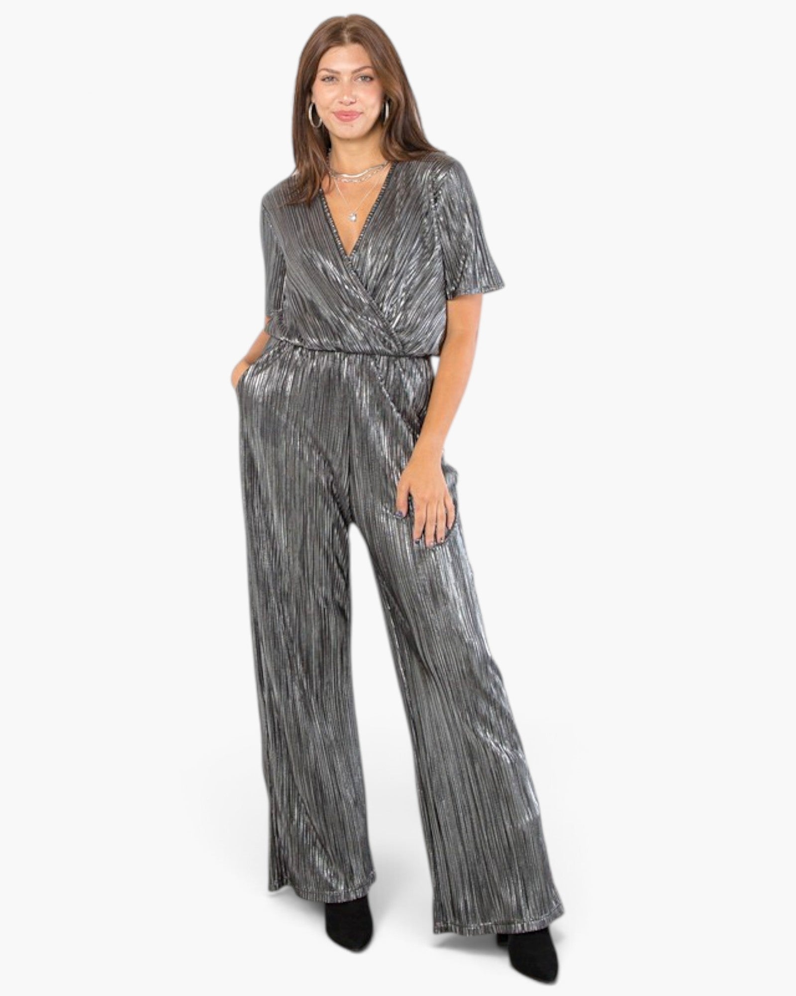 A woman modeling a metallic silver jumpsuit with a wrap-style V-neck bodice, short sleeves, and wide legs, photographed against a plain light gray studio background.