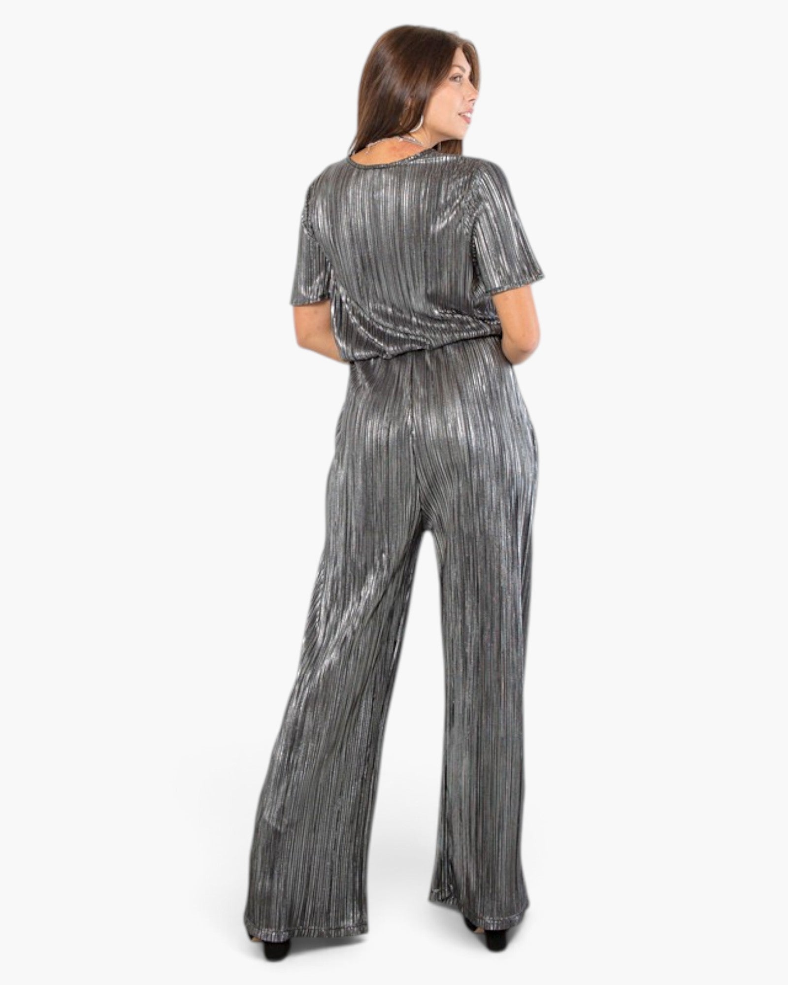 A woman modeling a metallic silver pleated jumpsuit from the back, featuring short sleeves and wide-leg pants, against a plain white background.