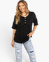 A woman modeling a black short-sleeve henley top with gold buttons, paired with distressed light-wash jeans, against a plain white studio background.