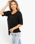 A woman modeling a black short-sleeve top with a subtle tonal pattern and a small button placket, paired with light-wash distressed jeans against a plain white background.