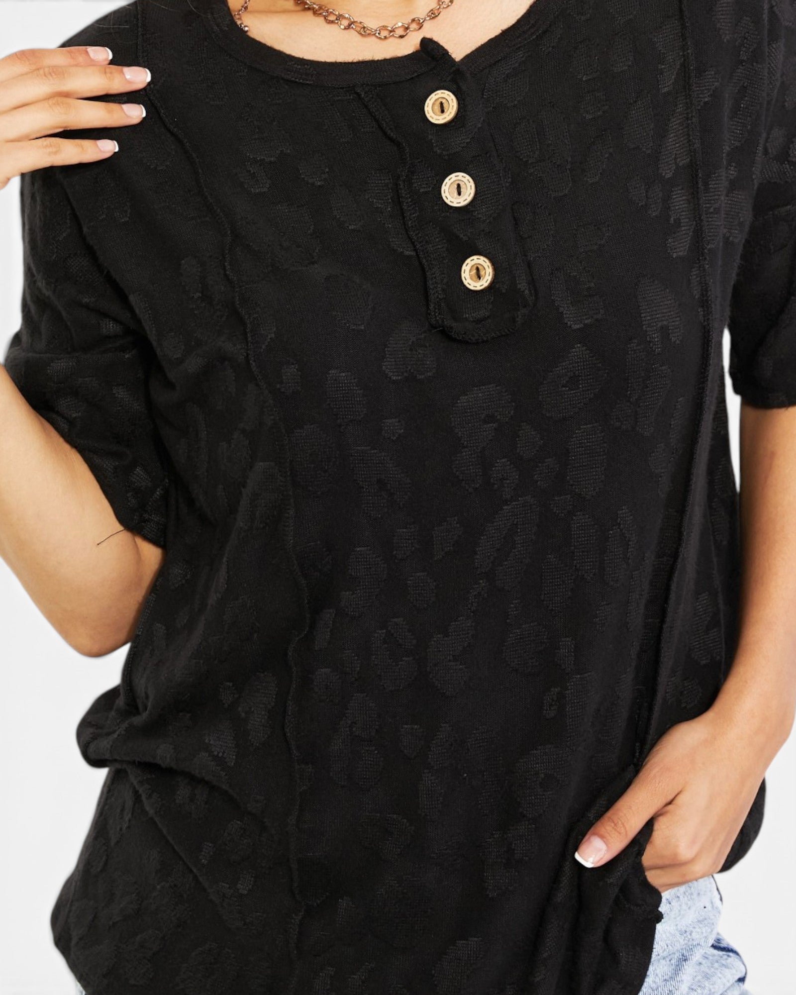 A close-up of a woman wearing a black short-sleeve Henley top with a subtle tonal leopard-like pattern and three gold buttons at the chest.