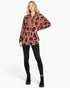 A woman modeling a long-sleeve button-up shirt with a black leopard print on a dusty rose background, paired with black skinny pants and black lace-up ankle boots, against a plain white studio background.