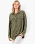 A woman modeling an oversized olive green textured button-up shirt with two chest pockets, paired with light-colored pants, against a plain white studio background.