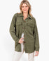 A woman modeling an olive green textured overshirt with brown buttons and flap chest pockets, paired with white pants, against a plain white background.