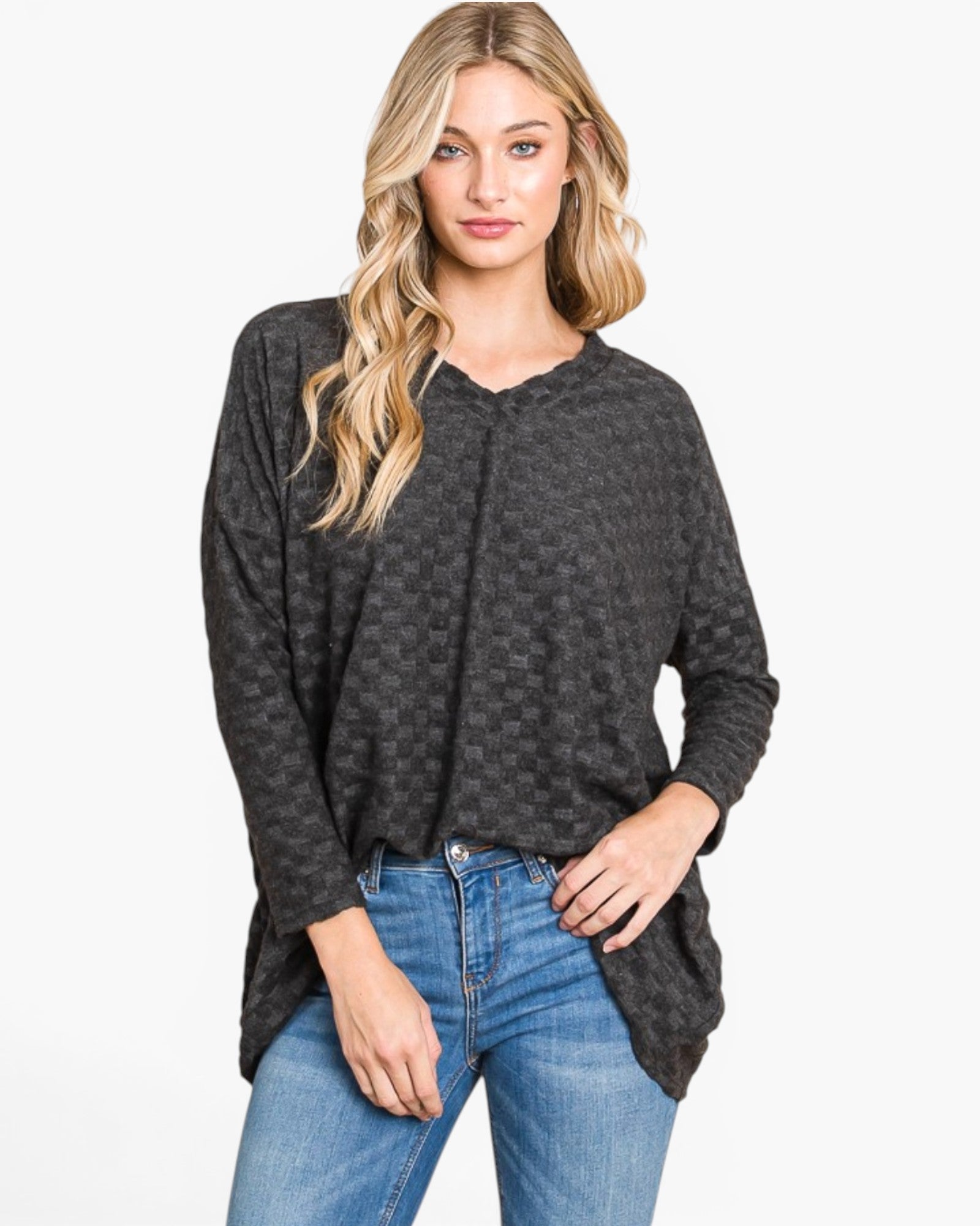A woman modeling a dark gray long-sleeve knit top with a subtle checkerboard pattern, paired with blue jeans, against a plain white studio background.