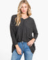 A woman modeling a dark gray long-sleeve knit top with a subtle checkerboard pattern, paired with blue jeans, against a plain white studio background.